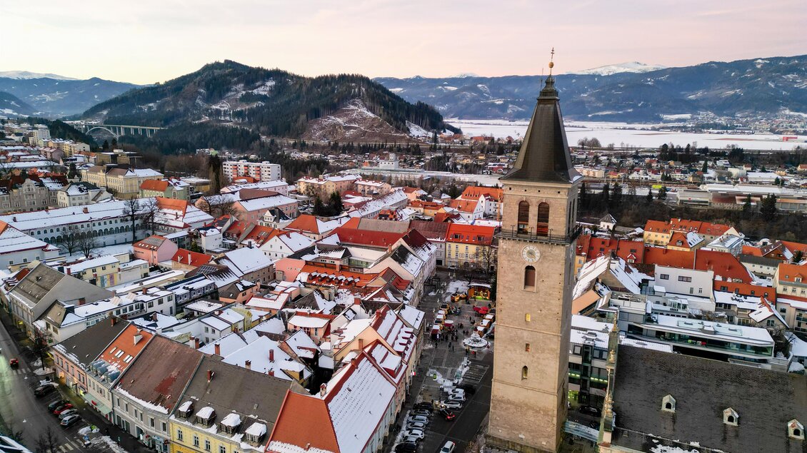 Eine malerische Stadtansicht mit schneebedeckten Dächern und einer markanten Kirchturmuhr. Im Hintergrund erkennt man sanfte Hügel und klaren Himmel. | © Isabella Painhapp