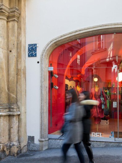 Ein Modegeschäft mit großem Schaufenster in leuchtendem Rot. Menschen gehen vorbei und betrachten die Auslage mit der Aufschrift „LAURA“. | © Graz Tourismus - Harry Schiffer