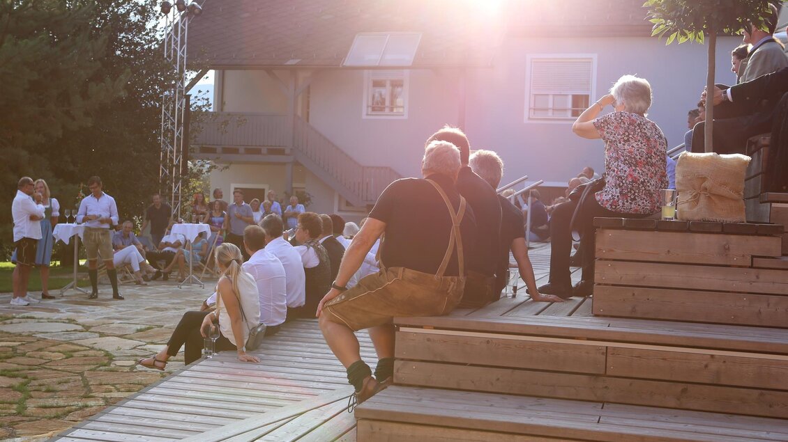 Eine Veranstaltung im Freien mit Menschen, die auf Holzbänken sitzen und einer Präsentation zuhören. Die Sonne scheint hell in eine entspannte Atmosphäre. | © Katharina Zotter