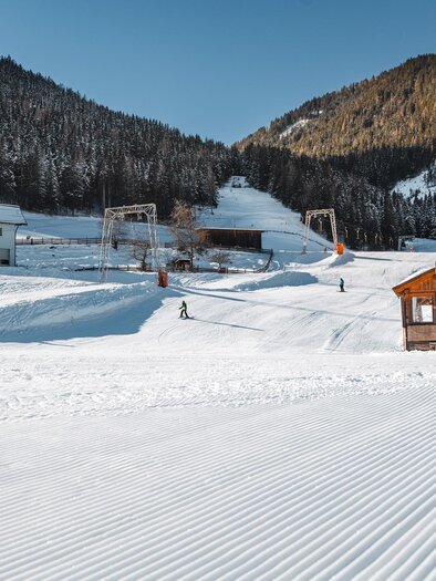 A snowy landscape with skiers and a small wooden house. The mountains in the background are surrounded by fir trees. | © Christoph Lukas