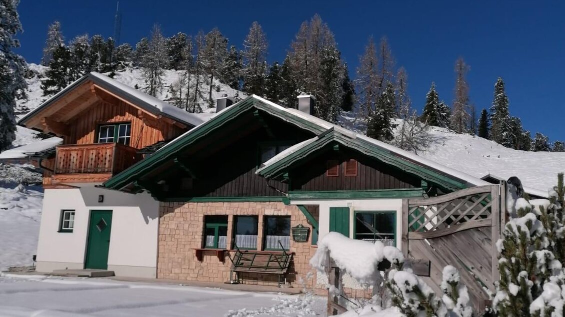 Ein gemütliches Haus im Schnee mit einem klaren blauen Himmel. Umgeben von verschneiten Bäumen und Bergen. | © Irmi Stieg