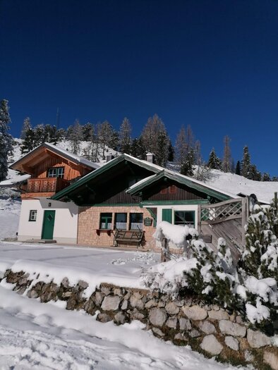 Ein gemütliches Haus im Schnee mit einem klaren blauen Himmel. Umgeben von verschneiten Bäumen und Bergen. | © Irmi Stieg