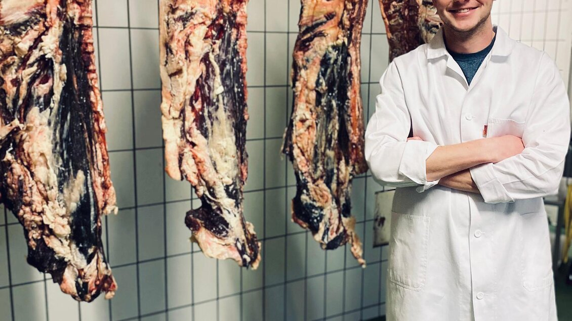 Ein junger Mann in einem weißen Arbeitsmantel steht in einer Fleischerei. Hinter ihm hängen mehrere Fleischstücke an Haken. | © Andreas Spanberger