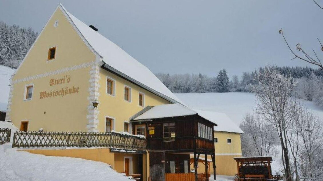 Ein charmantes gelbes Haus im Winter, umgeben von Schnee und schneebedeckten Bäumen. Auf der Veranda steht ein Holzpavillon, der einladend wirkt. | © Stoxreiter