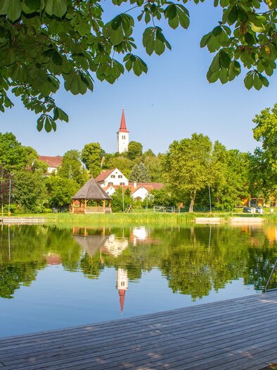 Ein ruhiger See umgeben von Bäumen und grünem Gras. Im Hintergrund steht eine kleine Kirche mit einem roten Turm. | © Sandra Brünner