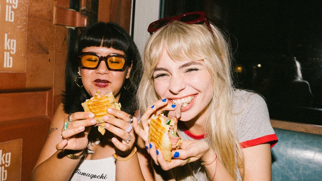  Zwei junge Frauen essen lachend Sandwiches in einem Sitzbereich bei Nacht, neben einem Container. | © Bobby's Agency - Simon Bauer
