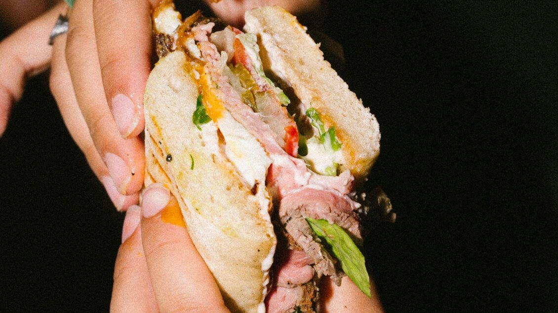 Nahaufnahme eines aufgeschnittenen Sandwichs mit Fleisch, Salat, Tomate, Sauce und Käse in Händen | © Bobby's Agency - Simon Bauer