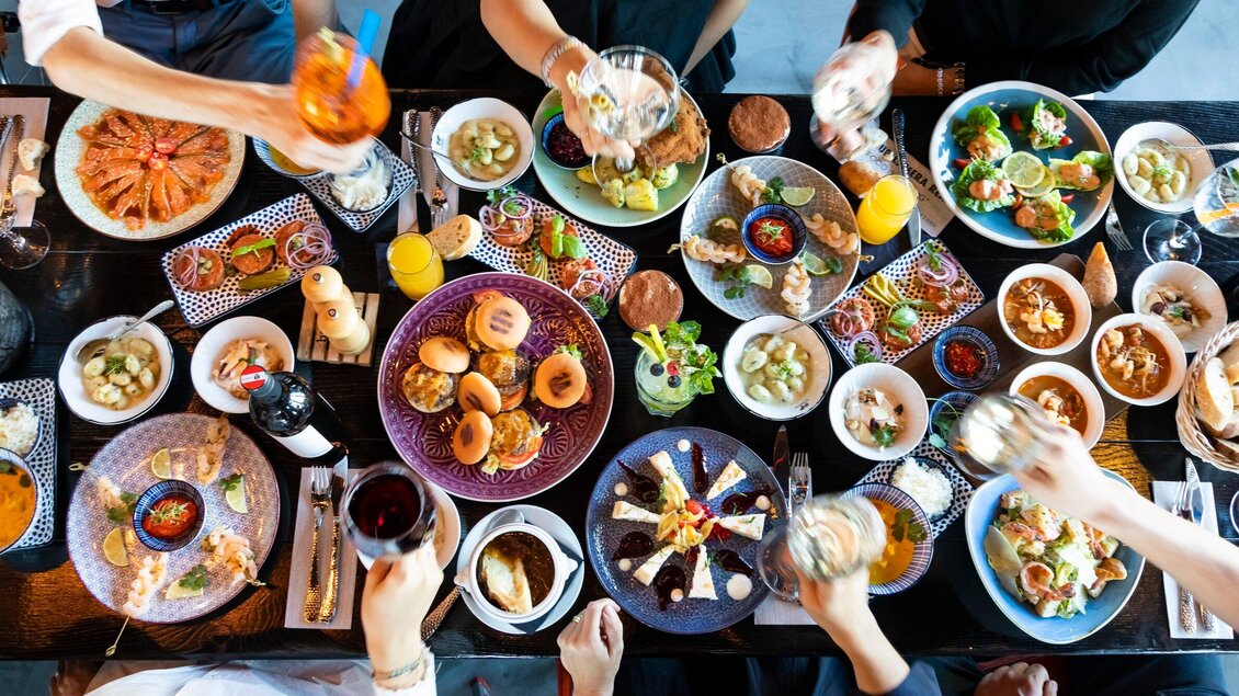Ein Tisch voller vielfältiger Gerichte und Getränke. Menschen stoßen mit Gläsern an und genießen ein festliches Essen. | © Streets - Werner Krug