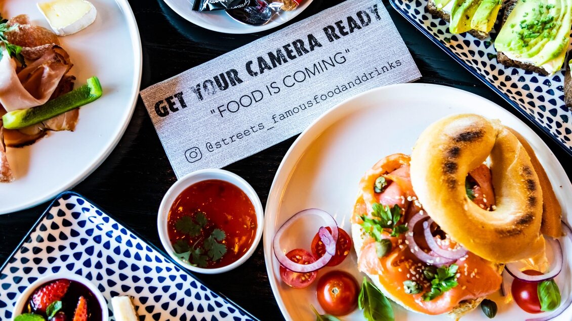  Frühstückstisch mit Bagel mit Lachs, Avocadobrot, Aufschnitt, Käse, Obst und Hinweis für Foto-Posting. | © Streets