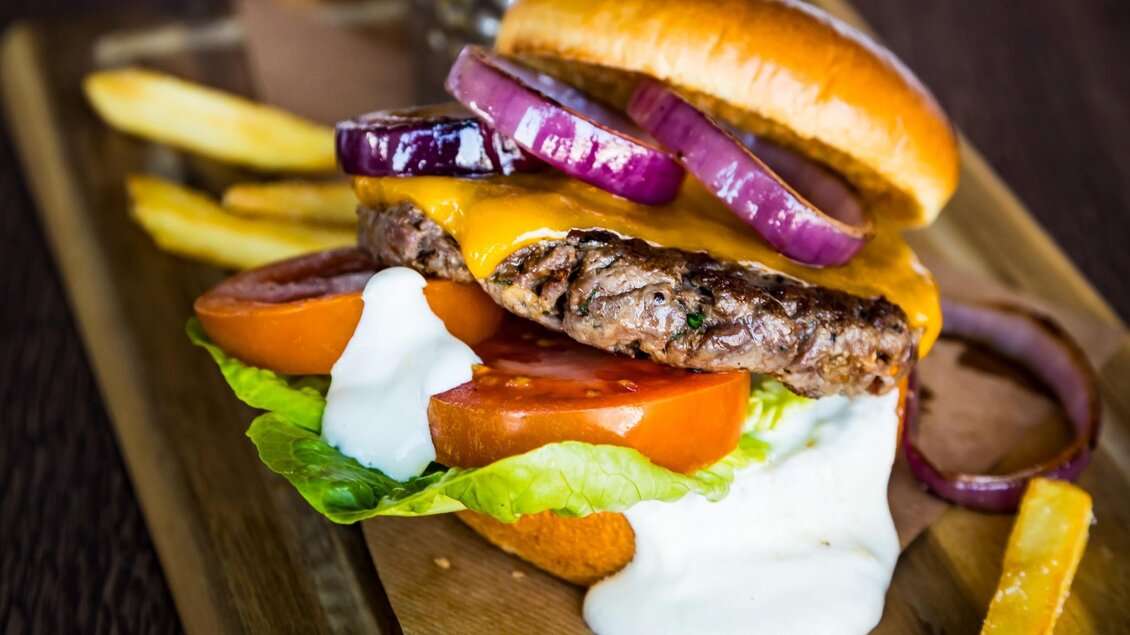  Nahaufnahme eines Burgers mit Cheddar, roten Zwiebeln, Tomate, Salat und Sauce, serviert mit Pommes | © Streets