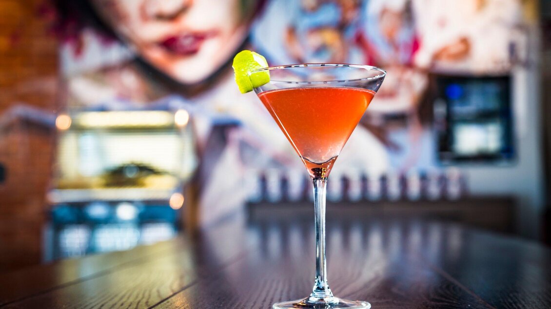 Ein eleganter Cocktail in einem Glas mit einem Stück Limette. Im Hintergrund ist ein farbenfrohes Wandgemälde zu sehen. | © Streets - Werner Krug