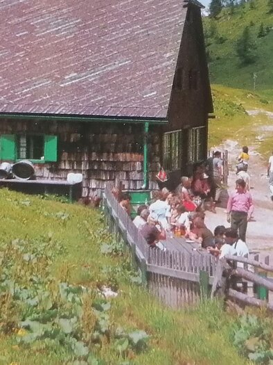 A picturesque landscape with traditional wooden houses. People gather outdoors, surrounded by green meadows and hills. | © Almgasthaus Göriacheralm / Familie Strobl