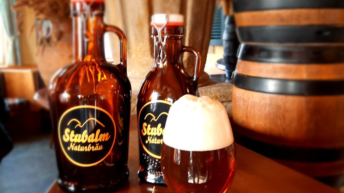 Zwei braune Bierflaschen und ein Glas mit schaumigem Bier stehen auf einem Tisch. Im Hintergrund sieht man ein großes Holzfass. | © Stubalmbräu-Mara
