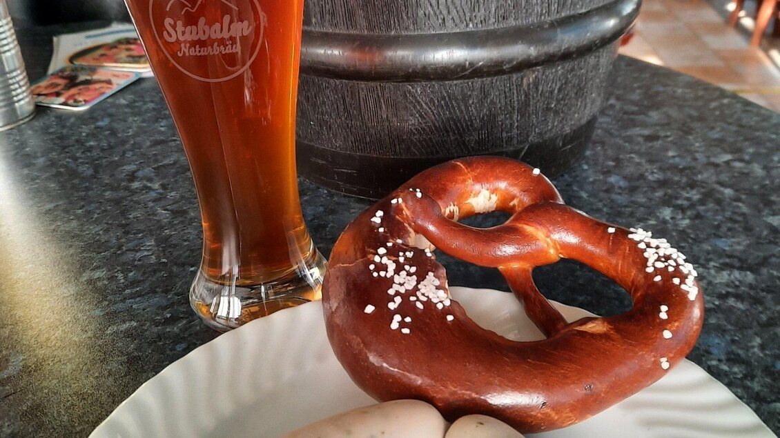 Ein frisch gebackenes Brezel auf einem Teller neben einigen Eiern und einem Glas dunklem Bier. Der Tisch hat eine rustikale Ausstattung. | © Stubalmbräu-Mara