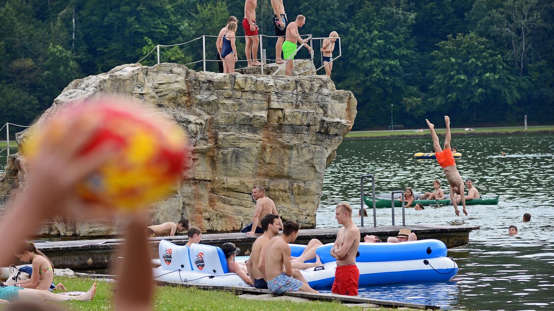 Eine Gruppe von Jugendlichen springt von einem Felsen ins Wasser. Am Ufer entspannen andere Badegäste auf Luftmatratzen und genießen den Sommertag. | © Tourismusverband Oststeiermark