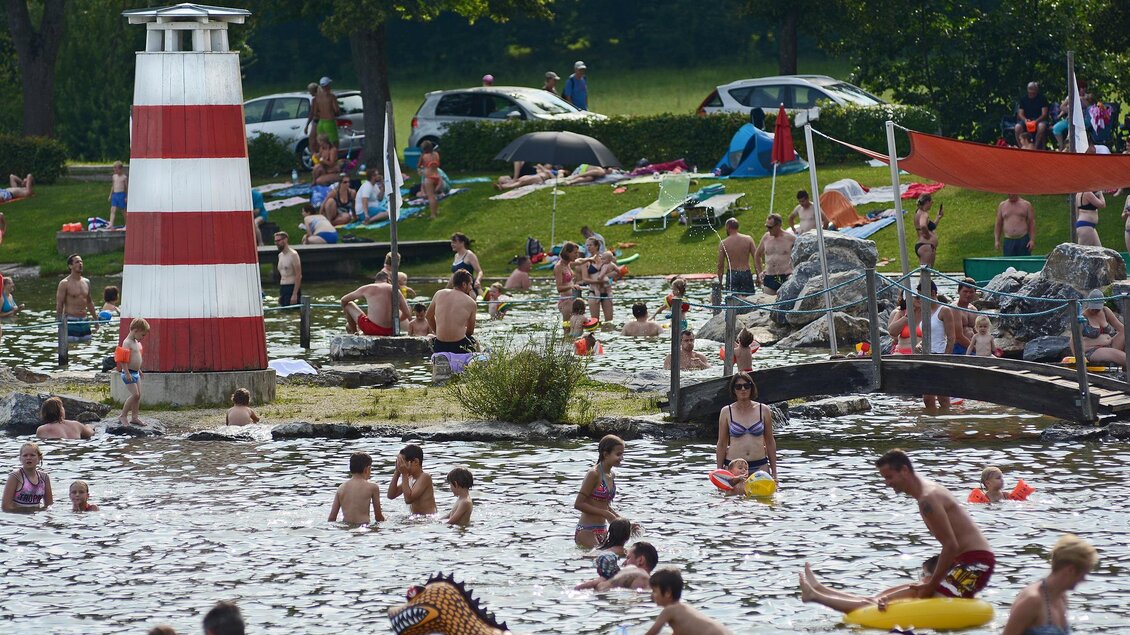 Ein belebter Badesee mit vielen Menschen. Kinder spielen im Wasser und es gibt einen roten und weißen Leuchtturm im Hintergrund. | © Tourismusverband Oststeiermark