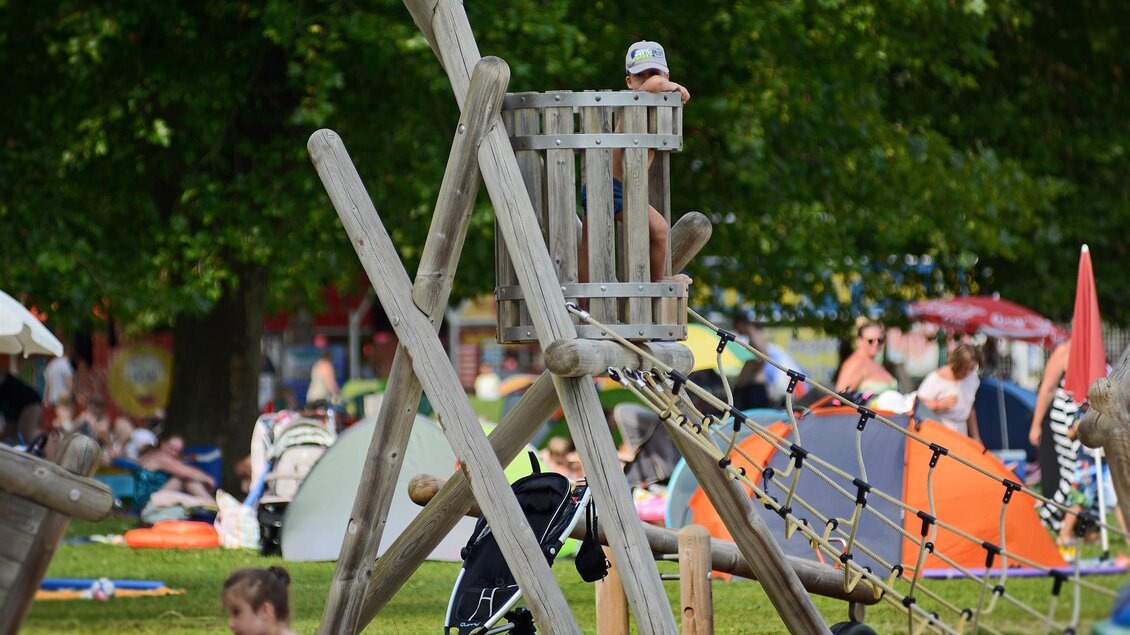 Ein Spielplatz mit einem Holzgerüst und einem Kletterseil. Im Hintergrund sind viele Menschen und Zelte zu sehen. | © Tourismusverband Oststeiermark