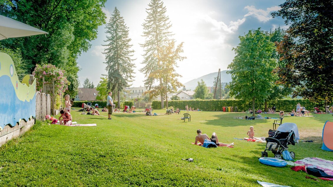 Die Liegewiese im Stukitzbad in Graz ist mit vielen Menschen belegt, die auf dem Gras entspannen. Im Hintergrund sind Bäume und Berge zu sehen. | © Holding Graz - Joel Kernasenko