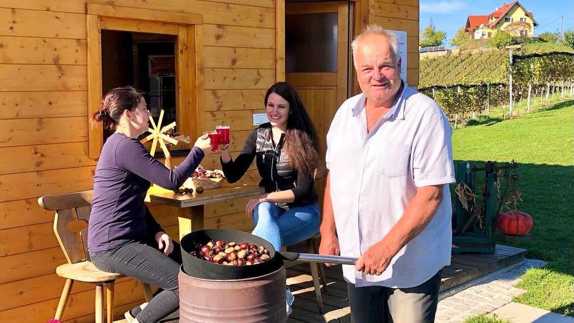 Eine gemütliche Holzlaube mit dem Schriftzug "Kraftplatzerl". Drei Personen genießen die Zeit draußen, während einer am Grill steht. | © Klapotetzhof