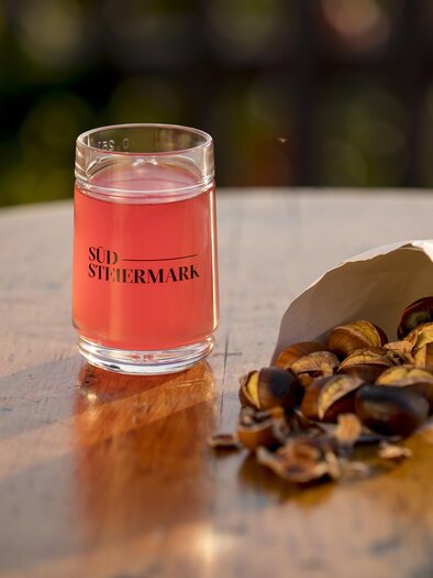 A glass with a pink drink is standing on a wooden table. Next to the glass lies a white paper bag with roasted chestnuts. | © Tom Lamm