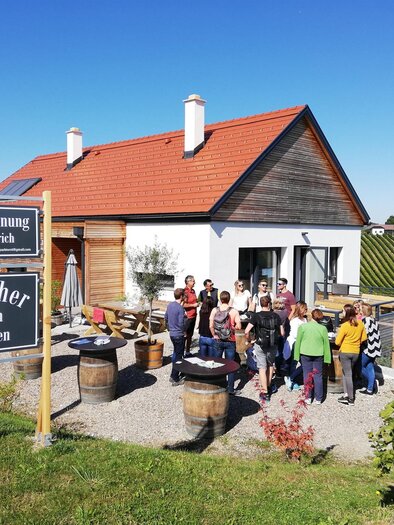 A modern house with a group of people outside. There are signs indicating a holiday apartment and a farm shop. | © Elke Ertl
