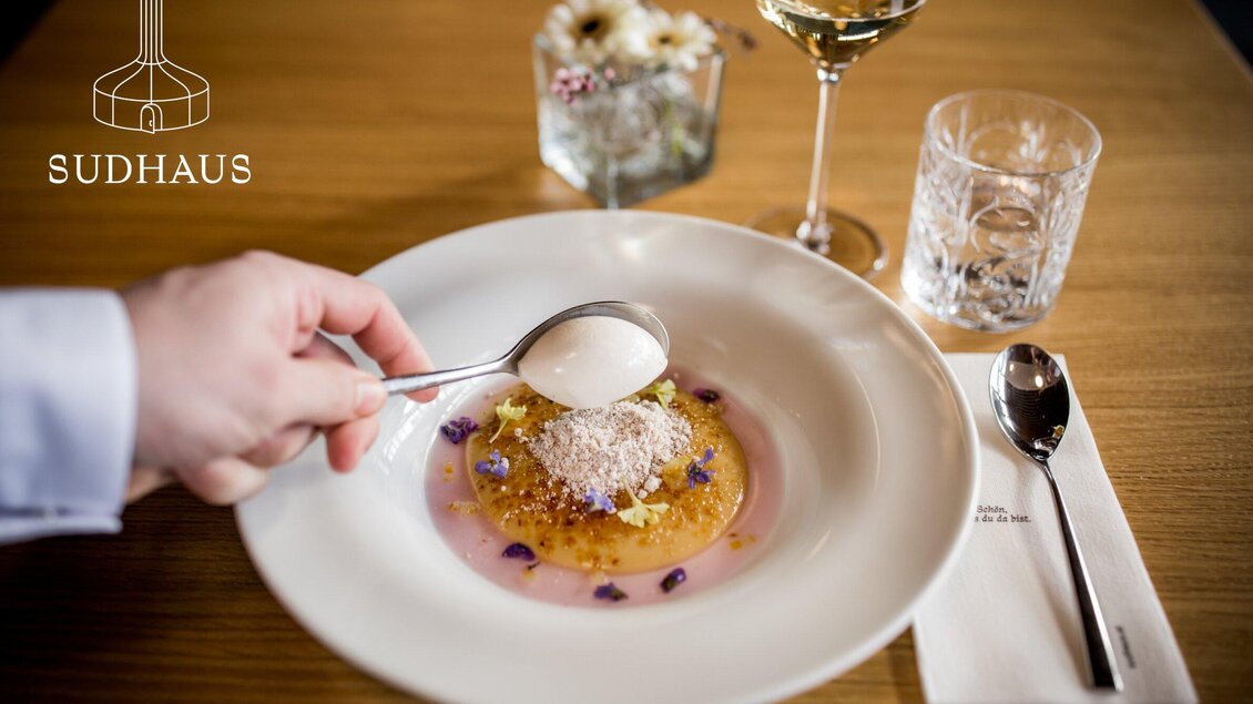 Ein elegantes Gericht wird serviert, mit einer Hand, die einen Löffel hebt. Im Hintergrund sind ein Glas Wein und ein Kristallglas sichtbar. | © Sudhaus