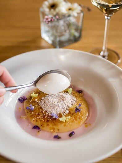 Ein elegantes Gericht wird serviert, mit einer Hand, die einen Löffel hebt. Im Hintergrund sind ein Glas Wein und ein Kristallglas sichtbar. | © Sudhaus