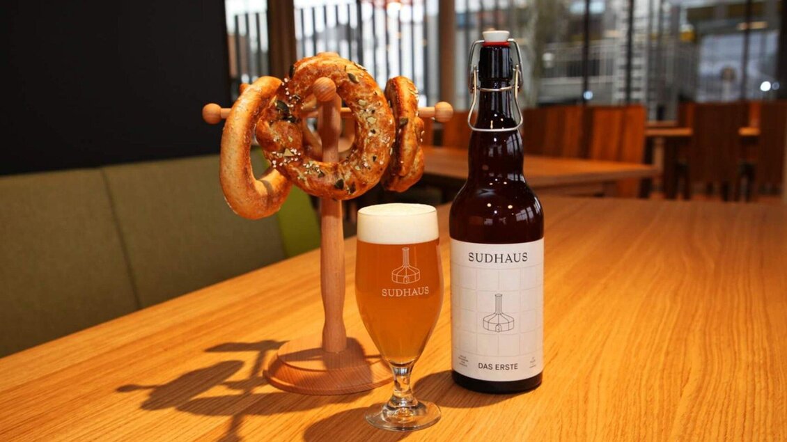 Ein Glas Bier und eine Flasche auf einem Tisch, daneben ein Halter mit Brezeln. Die Umgebung ist modern und gemütlich. | © Sudhaus