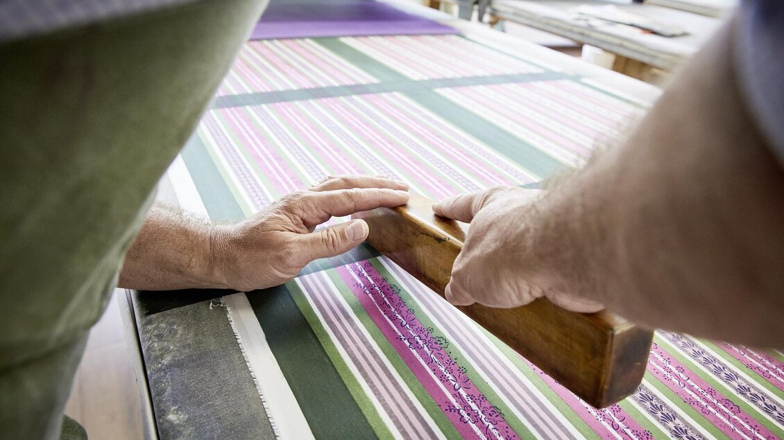 Ein Handwerker bearbeitet ein gestreiftes Stoffmuster mit einem Holzwerkzeug. Die Hände sind auf dem Tisch platziert, während sie präzise arbeiten. | © Handdrucke Sekyra-Helge Kirchberger