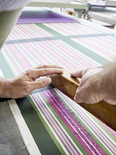 Ein Handwerker bearbeitet ein gestreiftes Stoffmuster mit einem Holzwerkzeug. Die Hände sind auf dem Tisch platziert, während sie präzise arbeiten. | © Handdrucke Sekyra-Helge Kirchberger