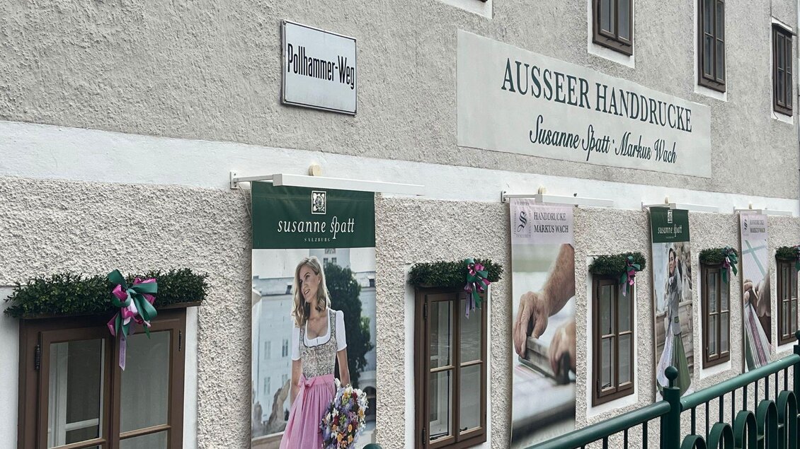 Eine Wand eines Gebäudes mit dem Schild "Ausseer Handdrücke" und mehreren Werbeplakaten. Die Plakate zeigen traditionelle Kleidung und schöne Blumenarrangements. | © TVB Ausseerland Salzkammergut_ Nathalie Schröttenhamer