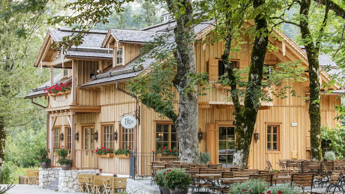 Ein charmantes Holzhaus umgeben von Bäumen. Draußen stehen Tische und Stühle in einem einladenden Garten. | © Seewiese Altaussee