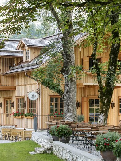 A charming wooden house surrounded by trees. Outside, tables and chairs are set up in an inviting garden. | © Seewiese Altaussee