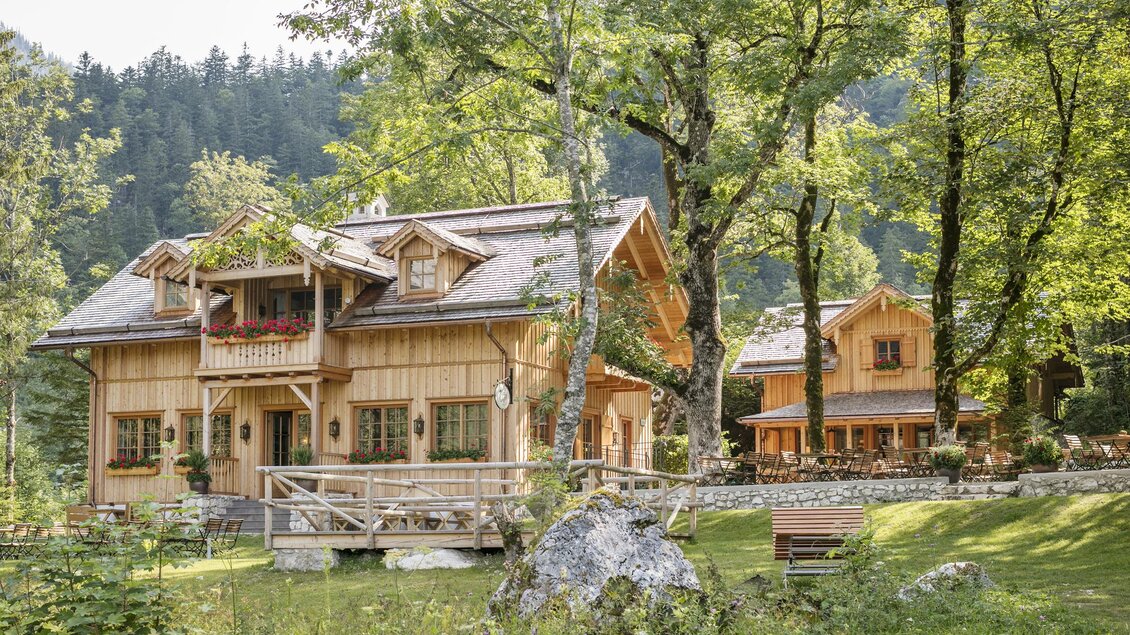 Ein malerisches Holzhaus umgeben von Bäumen und grüner Landschaft. Im Hintergrund sind weitere Gebäude und sanfte Hügel zu sehen. | © Seewiese Altaussee