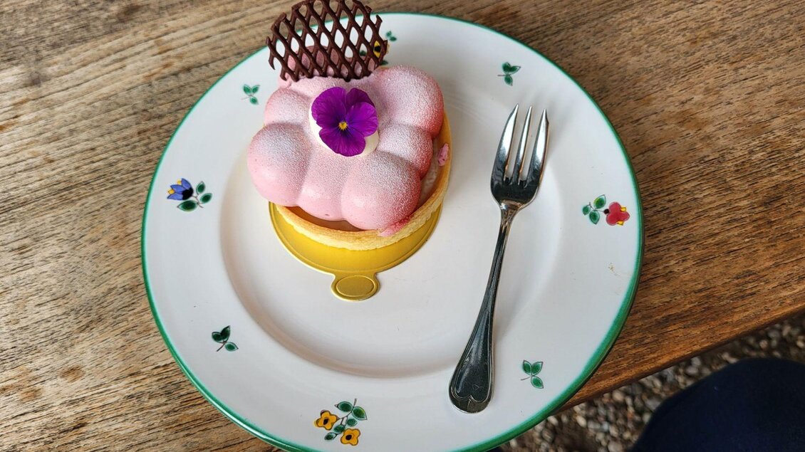 Ein kunstvoller Dessertteller mit einem rosa Kuchen, dekoriert mit einer Blume und einem Schokoladennetz. Der Kuchen liegt auf einem festlichen Teller mit Blumenmotiven. | © Petra Kirchschlager