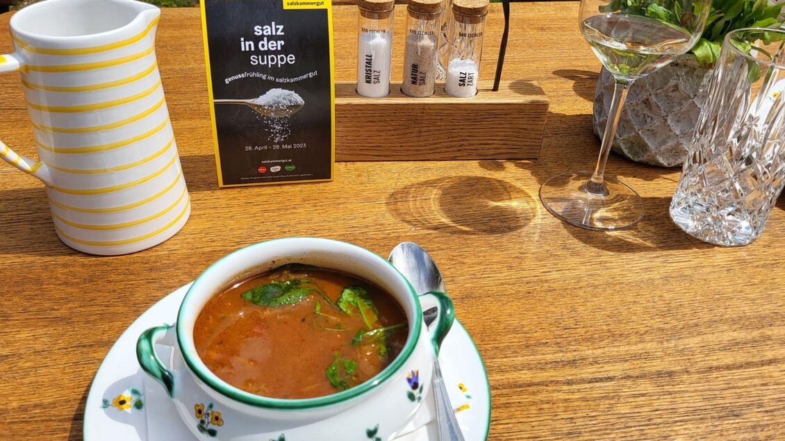 Eine Schüssel Suppe mit frischen Kräutern steht auf einem Holztisch. Im Hintergrund sind ein Wasserglas, eine Karaffe und ein Schild mit dem Text „Salz in der Suppe“ zu sehen. | © Petra Kirchschlager