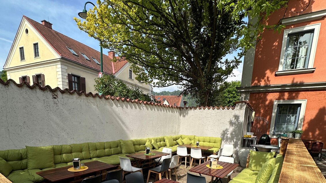 Eine einladende Terrasse mit grünen Sitzgelegenheiten und Holztischen. Im Hintergrund sind Bäume und bunte Gebäude zu sehen. | © TREND