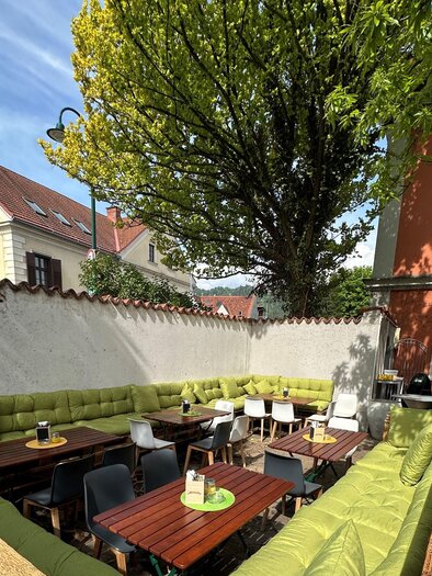 Eine einladende Terrasse mit grünen Sitzgelegenheiten und Holztischen. Im Hintergrund sind Bäume und bunte Gebäude zu sehen. | © TREND