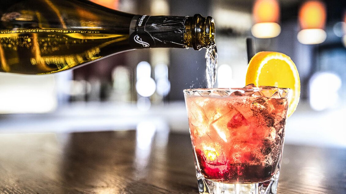 Ein Drink mit Eiswürfeln und einer Scheibe Orange wird eingeschenkt. Im Hintergrund sind unscharfe Lichter zu sehen. | © TUI BLUE