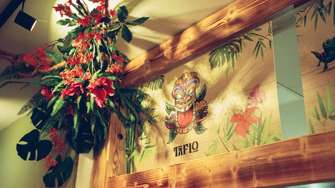 Ein schöner Innenraum mit tropischen Pflanzen und bunten Blumen. An der Wand ist ein farbenfrohes Gemälde mit einem Maskenmotiv zu sehen. | © Julian Kroissenbrunner