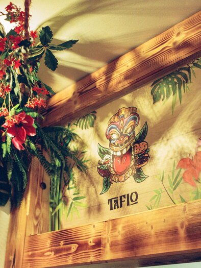 Ein schöner Innenraum mit tropischen Pflanzen und bunten Blumen. An der Wand ist ein farbenfrohes Gemälde mit einem Maskenmotiv zu sehen. | © Julian Kroissenbrunner