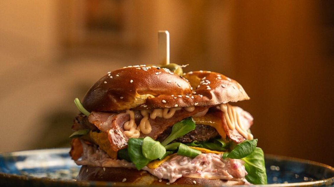 Ein leckerer Burger auf einem blauen Teller, garniert mit frischem Gemüse und einer Sauce. Im Hintergrund sind verschwommene Lichter zu sehen, die eine gemütliche Atmosphäre schaffen. | © Julian Kroissenbrunner