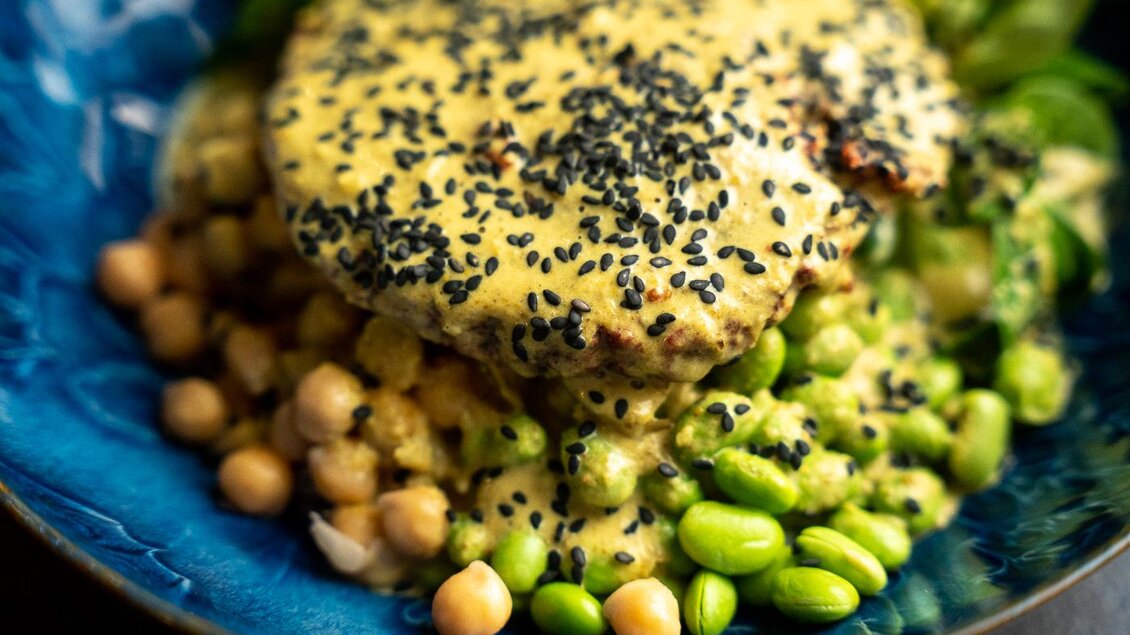 Ein bunter Teller mit Kichererbsen, grünen Sojabohnen und frischem Blattgemüse. Darüber liegt eine cremige Sauce mit schwarzem Sesam.