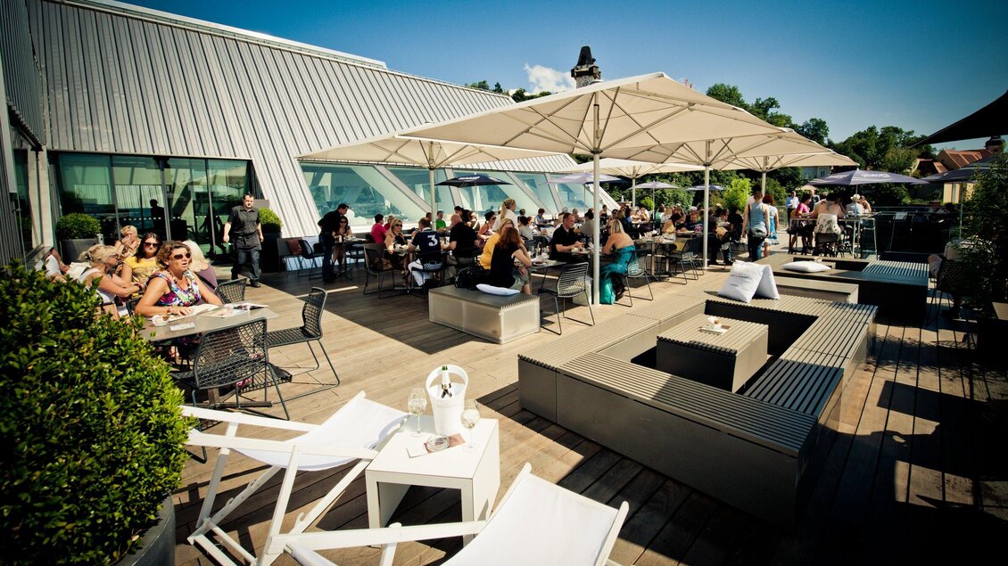 Eine belebte Dachterrasse mit vielen Gästen, die an Tischen sitzen. Schirme spenden Schatten, und die Atmosphäre ist gemütlich und einladend. | © K&Ö - Lupispuma