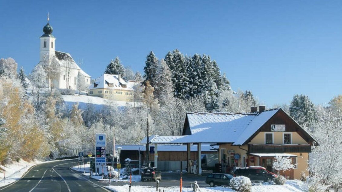 Eine verschneite Landschaft mit einer Straße, einem Bauernhaus und einer Kirche im Hintergrund. Der Himmel ist klar und blau. | © Tankstellenbuffet Golob