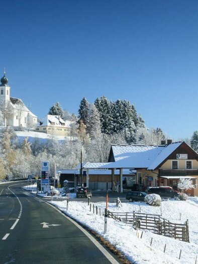 A snowy landscape with a road, a farmhouse, and a church in the background. The sky is clear and blue. | © Tankstellenbuffet Golob
