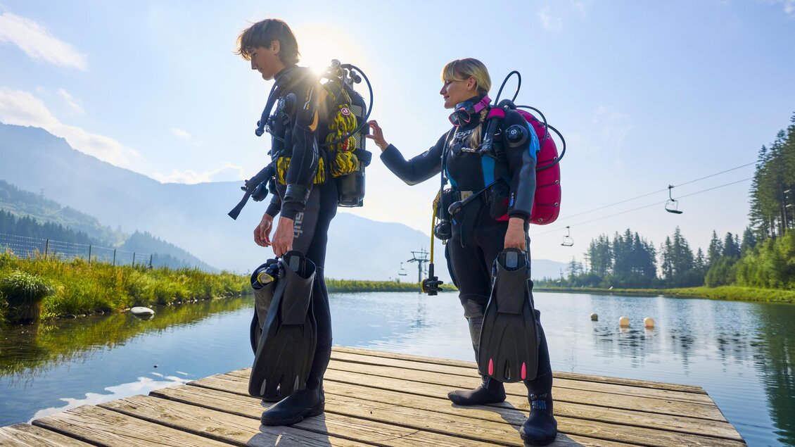 Zwei Taucher stehen auf einem Holzsteg am Wasser. Die Sonne scheint und die Landschaft ist von Bergen und Bäumen umgeben. | © Susanne Einzenberger