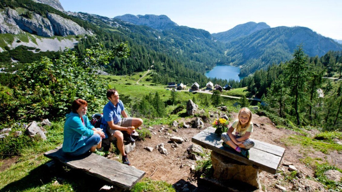 Eine malerische Berglandschaft mit einem klaren See im Hintergrund. Eine Familie sitzt auf einer Holzbank und genießt die Aussicht. | © TVB Ausseerland Salzkammergut_Tom Lamm
