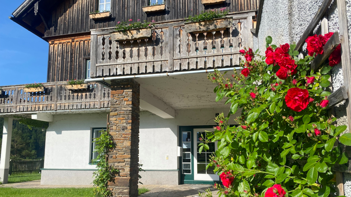 Ein charmantes Haus mit Holzfassade und einem Balkon voller Blumen. Vor dem Haus blühen rote Rosen und die Sonne scheint auf das Grundstück. | © Bettina Scheck
