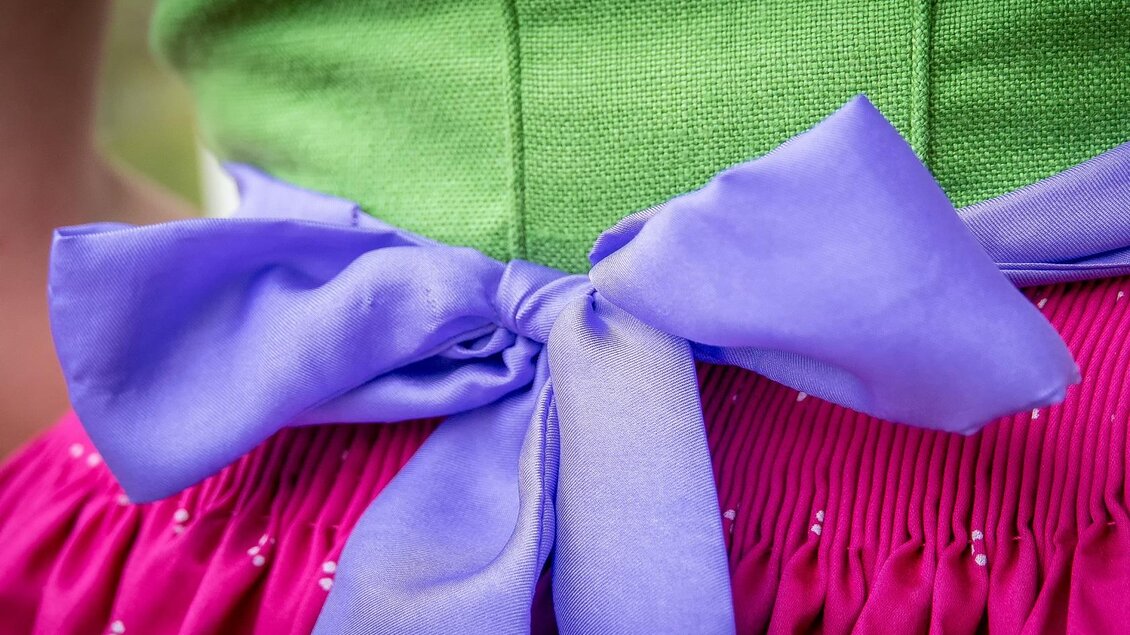 Ein detailreicher Ausschnitt eines Kleides mit einer grünen Oberseite und einem pinken Rock. Der Fokus liegt auf einer lila Schleife, die den Rock betont. | © Christine Struz